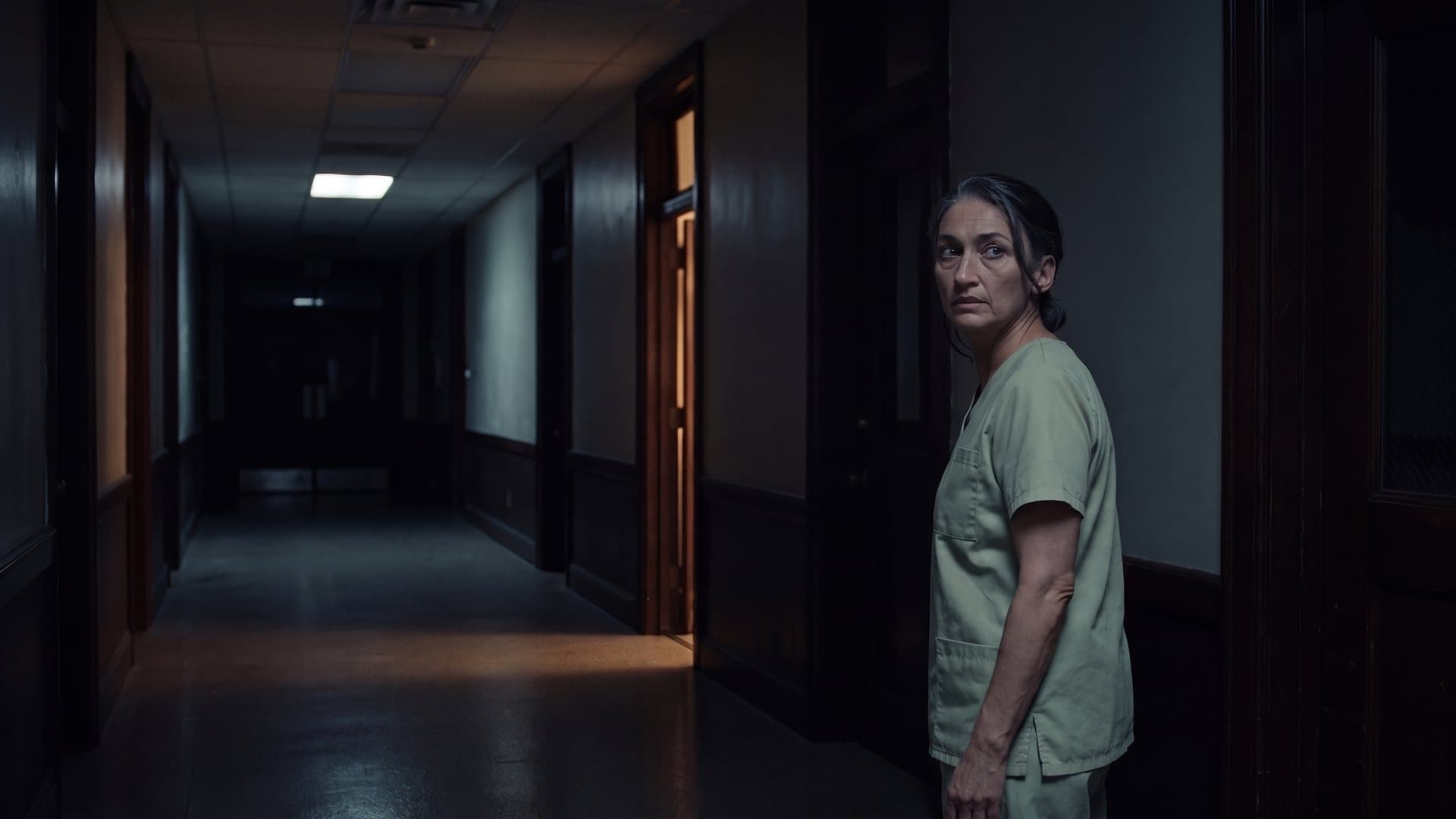 An orderly stops in a dark corridor beside a lit doorway and looks back over her shoulder.