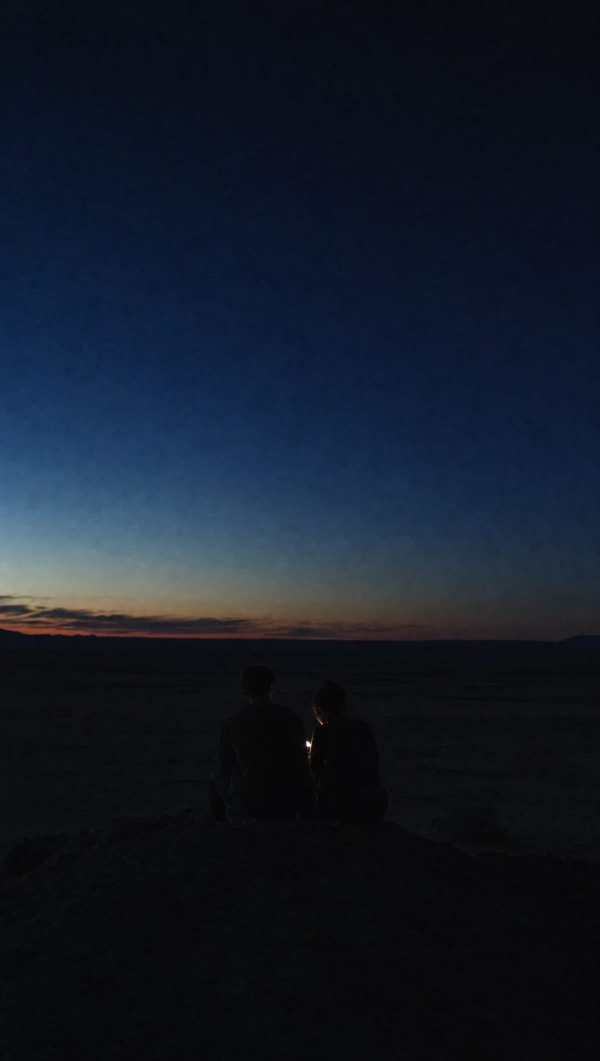 Two people sit together on a rock at deep dusk over a vast dark plain, a single small light glowing between them.